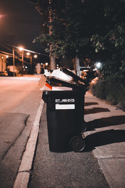 A black refuse bin with a white label and the text 'St. John's' positioned on a pavement along a dimly lit urban street scene at night. The lid of the bin is open, revealing a variety of discarded waste materials such as cardboard, paper, and plastic items. The bin is situated next to a curb, with the street illuminated by nearby streetlights casting warm light and shadows. In the background, blurred outlines of trees and street furniture are visible, contributing to a quiet, residential environment. The scene suggests an instance of independent waste disposal or rubbish collection outside a home or community area, with the focus on efficient rubbish management and disposal, relevant to services offered by companies providing private waste handling solutions in the local area.