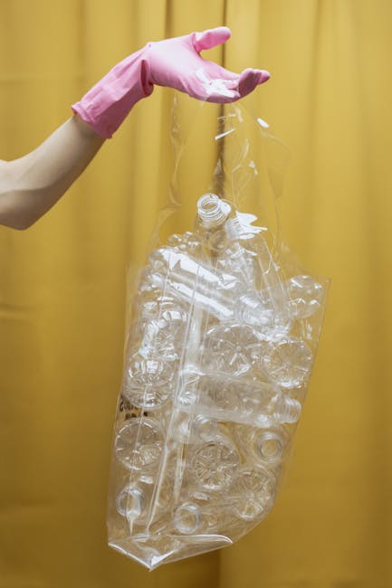A person wearing pink rubber gloves is holding a large, transparent plastic bag filled with discarded plastic bottles, rings, and containers, which are crumpled and partially crushed. The background features a plain, yellow curtain with soft lighting, giving a neutral, indoor environment. The scene emphasizes the collection and disposal of plastic waste, aligning with the context of rubbish removal services. The plastic waste appears clean and well-contained in the bag, illustrating proper handling of recyclable materials and supporting the concept of alternative waste management methods in residential or commercial settings. The focus is on the hand and the plastic bag, with the texture of the clear plastic and the various shapes of the bottles and containers clearly visible, reflecting the importance of proper waste segregation and disposal options provided by companies like rubbishcollectionhollandpark.co.uk.
