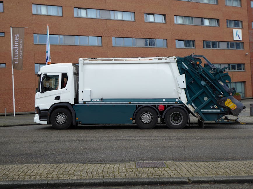 A large, modern waste collection vehicle parked parallel to a paved street in front of a multi-storey brick building with reflective windows. The truck features a white cab with black trim and a dark blue lower section, with a hydraulic lift or compactor mechanism at the rear, designed for efficient rubbish collection. The rear component, painted in matching dark blue, contains metal parts and hydraulic arms used for loading and compacting waste, with the yellow safety guard clearly visible. The vehicle is positioned on a curbside with a small strip of patterned paving alongside and a narrow sidewalk. In the background, the brick building has a horizontal row of narrow, rectangular windows and a signage pole with a vertical banner on the left side. The scene is illuminated by natural daylight, with no people visible, emphasizing the vehicle's role in independent waste handling outside typical municipal rubbish collection settings.