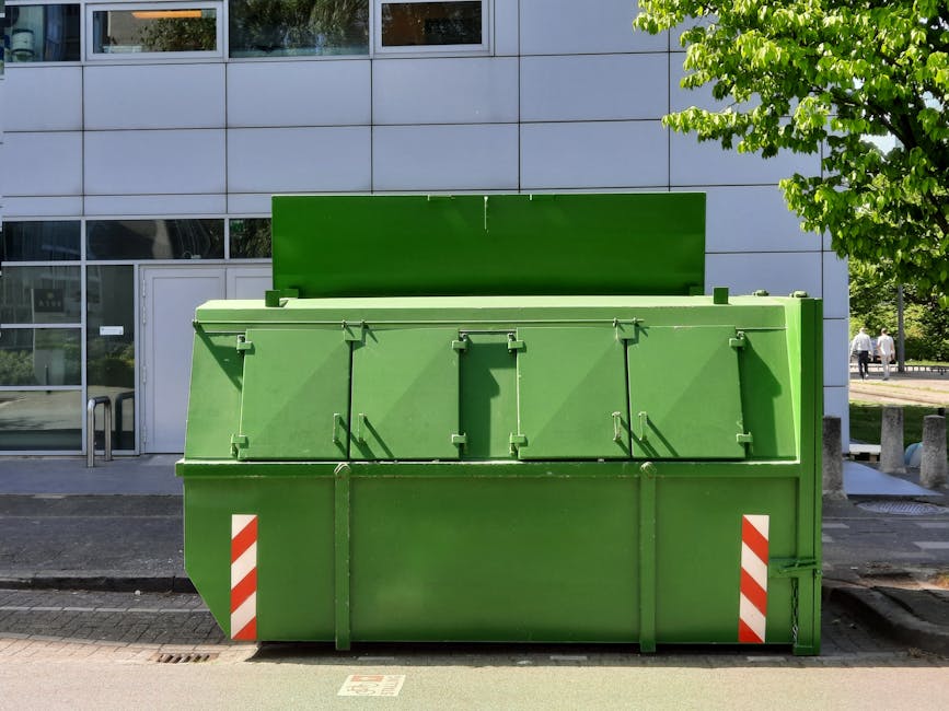 A large, commercial green waste container with a sturdy, rectangular metal structure and a smooth, flat lid positioned upright at the back, located on a paved sidewalk in front of a modern, grey institutional building with a glass entrance. The container's surface has a matte finish, and it features small, latch-like mechanisms on the front for securing the lid, along with diagonal red and white reflective stripes on the bottom corners to enhance visibility. Surrounding the container are elements of urban infrastructure, including a sloped curb and a metal bike rack to the left, with a tree and some pedestrians visible in the background. The building's façade is composed of large, square panels, and the scene is illuminated by bright daylight, casting shadows on the container and the nearby pavement. This setup suggests an organized location for private waste collection, reflective of alternative rubbish disposal solutions outside conventional council services, as highlighted by Holland Park waste collection practices.