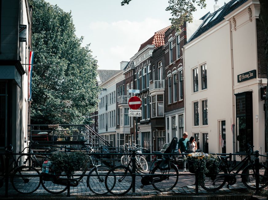 An urban street scene in a residential area features a row of multi-storey buildings with a mix of white and dark brick facades, some with decorative window trim and small balconies. In the foreground, several bicycles are securely locked to black metal bike racks along the paved sidewalk. Two pedestrians, a man and a woman, are walking near the corner, engaged in conversation. A large tree on the left side provides partial shade, its branches extending over part of the scene. A street sign indicating a no-entry zone for vehicles is mounted on a pole behind the pedestrians, with smaller directional and bicycle route signs beneath it. The overall environment reflects a typical city centre with closely spaced buildings, outdoor fixtures, and a calm atmosphere, subtly connecting to the theme of alternative waste handling or private rubbish collection in urban neighbourhoods, as visual cues suggest a well-maintained, residential community setting.
