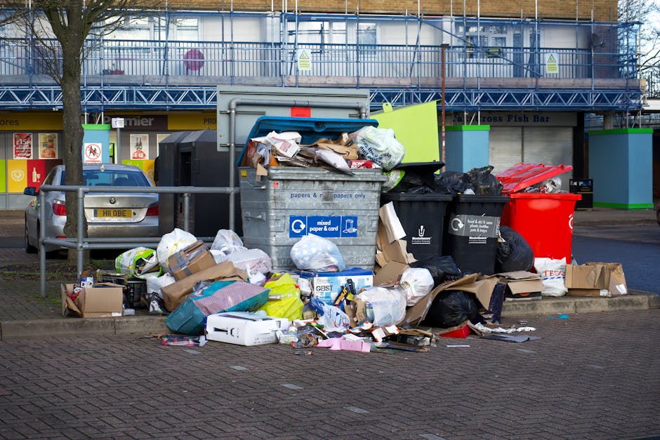 A large overflow of mixed waste, including crumpled cardboard boxes, plastic bags, paper, and packaging materials, is scattered on a paved sidewalk beside a metal railing. Behind the waste, several black, red, and yellow wheelie bins are visible, with some lids open and contents spilling out. A silver hatchback car is parked nearby on the left side of the image, adjacent to a row of shopfronts that feature signage and storefront displays. In the background, a building under renovation is covered with blue scaffolding and protective netting, indicating ongoing construction work. The entire scene depicts a typical rubbish accumulation in an urban area, illustrating the potential consequences of informal waste disposal or fly-tipping, and highlighting the need for proper waste management and alternative rubbish collection services such as those provided by Holland Park's waste specialists, including companies like rubbishcollectionhollandpark.co.uk. The environment appears damp, with overcast lighting, emphasizing the cluttered and unmanaged state of the waste scene.