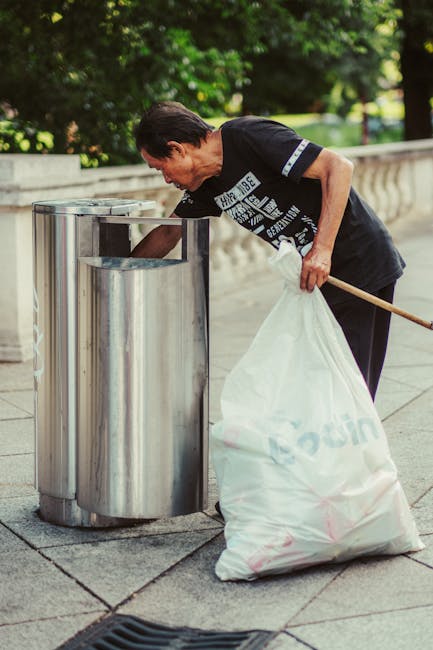 A middle-aged man with dark hair, dressed in a black T-shirt with white graphic text, is bent over placing waste into a silver, cylindrical metal rubbish bin situated on a paved sidewalk. He is holding a large white plastic bag filled with rubbish in his left hand, resting against his side, while his right hand reaches towards the opening of the bin. The bin has a rounded top with an opening for waste disposal, and the surface reflects the surrounding environment. Behind him, there is a low stone balustrade which runs parallel to the sidewalk, and lush green foliage of trees and shrubs provides a natural backdrop, indicating an outdoor public space. The scene is illuminated by natural sunlight, creating a clear and bright atmosphere, and the overall setting suggests a typical urban or park environment focused on waste management or rubbish collection, possibly related to private or community rubbish clearance services as seen in similar contexts involving rubbish removal in Holland Park. The scene emphasizes practical waste disposal in a clean, well-maintained public area, consistent with alternative or independent rubbish collection methods.