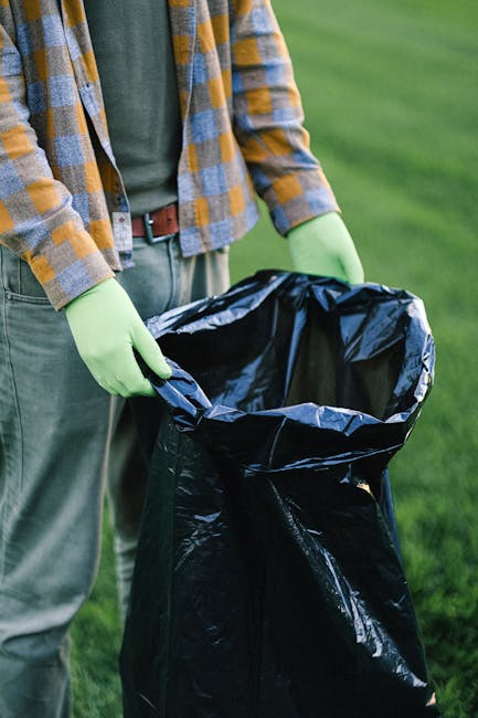 A large outdoor waste collection point featuring multiple open-top wheelie bins and numerous overflowing rubbish bags, cardboard boxes, and loose paper scattered around the base. The central bin is made of grey plastic with a blue recycling symbol and the label 'mixed paper & card,' overflowing with crumpled newspapers, cardboard cartons, and paper waste. To its right, there are black and red bins, both filled with various waste items, with some debris spilling onto the surrounding pavement. In front of the bins, an assortment of waste, including flattened cardboard boxes, plastic bags, tissue paper, and packaging materials, are strewn across the brick-paved ground, indicative of an on-site rubbish disposal situation. Behind the waste area, a street scene is visible with parked cars, a tree with bare branches, and a building façade with storefront signs and a blue scaffolding structure, suggesting a commercial or residential area undergoing maintenance. The scene reflects a typical scene that might occur in independent rubbish collection or private waste disposal contexts in urban environments, which waste management services like those in Holland Park might handle or monitor.