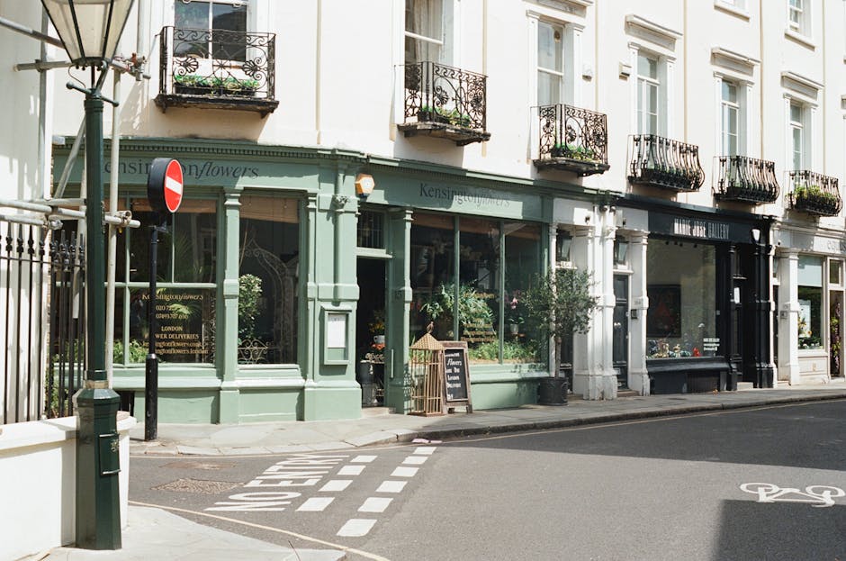 A daytime street view showing a row of commercial buildings with large glass display windows and decorative black wrought-iron balconies on the upper floors. The building on the left, painted in a pastel green shade, has a flower shop with potted plants and floral arrangements visible through the windows. A blackboard sign is placed outside the shop, and a small cage with a bird decoration leans against the window. Adjacent buildings are painted in light neutral tones, with some having similar iron balconies and potted plants. A narrow pavement runs alongside the buildings, with a street lamp and a lamppost situated on the sidewalk. In the foreground, a bicycle lane is marked on the asphalt, with a treaty signpost indicating no entry for vehicles from the left. The scene is well-lit, with natural daylight casting soft shadows, emphasizing the clean, orderly appearance of the storefronts and urban environment, characteristic of independent shop access and pedestrian-friendly areas in a city setting.