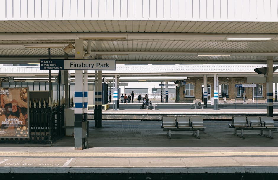 The image depicts Finsbury Park train station platform, with a concrete canopy roof overhead supported by metal columns. A white sign with black lettering reading 'Finsbury Park' is centrally positioned, hanging below the roof. Several passengers are seated on metal benches, some waiting, while others are walking on the platform or approaching the adjacent tracks. The platform surface is made of textured concrete with tactile paving strips near the edge for safety. In the background, additional platforms and a station building with brickwork, windows, and a small shelter are visible. To the left, a black fence with gold-colored spiked decoration runs along the platform boundary, next to a vibrant poster advertising a theatrical performance of 'Romeo and Juliet.' The scene is illuminated by natural daylight, with soft shadows cast on the platform, typical of a clear day. The visual context aligns with independent or private transport arrangements, relevant to rubbish removal services that may require site access to manage waste outside standard public disposal options, subtly connecting to the themes of alternative waste handling or on-site clearance seen in rubbish collection near urban transit hubs.