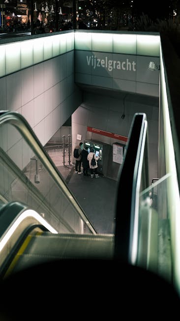 The image shows a view from the top of an escalator descending into the Vilzelgracht underground station. The station entrance is characterized by large, illuminated greenish-white panels forming the upper part of the walls. The descending escalator has metallic handrails and steps, with the metal surfaces reflecting ambient light. At the bottom of the escalator, there is a small group of three people standing in front of a red informational sign attached to a tiled wall. The tiled area features beige and light brown square tiles. Adjacent to the station entrance, there is a set of staircase railings and additional signage. The overall environment appears clean and modern, with the lighting suggesting artificial illumination typical of underground transit stations, supporting efficient passenger movement and safety. Although the scene is focused on public transportation, the crisp detail of the station’s architectural elements and station signage aligns with the context of alternative urban transportation solutions, similar to private disposal or on-site clearance services provided by companies like Rubbish Collection Holland Park.