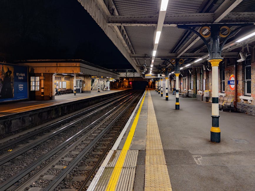 The image depicts Finsbury Park train station platform, with a concrete canopy roof overhead supported by metal columns. A white sign with black lettering reading 'Finsbury Park' is centrally positioned, hanging below the roof. Several passengers are seated on metal benches, some waiting, while others are walking on the platform or approaching the adjacent tracks. The platform surface is made of textured concrete with tactile paving strips near the edge for safety. In the background, additional platforms and a station building with brickwork, windows, and a small shelter are visible. To the left, a black fence with gold-colored spiked decoration runs along the platform boundary, next to a vibrant poster advertising a theatrical performance of 'Romeo and Juliet.' The scene is illuminated by natural daylight, with soft shadows cast on the platform, typical of a clear day. The visual context aligns with independent or private transport arrangements, relevant to rubbish removal services that may require site access to manage waste outside standard public disposal options, subtly connecting to the themes of alternative waste handling or on-site clearance seen in rubbish collection near urban transit hubs.