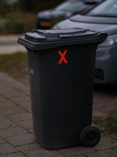 A middle-aged man with dark hair, wearing a black T-shirt with white graphics and dark trousers, is seen in an outdoor urban environment. He is bent over, placing a white plastic bag filled with rubbish into a metal public rubbish bin that has a cylindrical shape and a polished, reflective surface. The bin features a rectangular slot at the top for waste disposal and is positioned on a paved sidewalk. In the background, there is a stone balustrade and lush green trees, indicating a park or pedestrian area. The man is holding a wooden litter picker in his left hand while leaning forward to deposit the rubbish, demonstrating public waste disposal activity. The setting appears well-lit with natural daylight, and the overall scene is neutral and observational, capturing a typical moment of rubbish collection or disposal in a city environment, suitable for illustrating municipal or private waste handling services like those offered by rubbishcollectionhollandpark.co.uk.