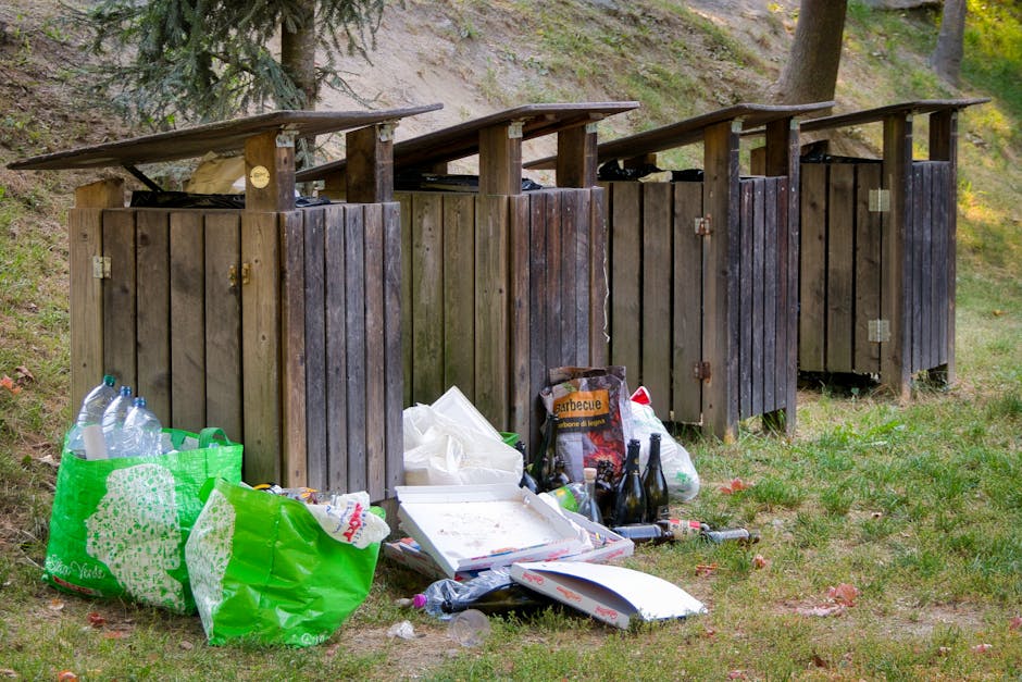 A series of four wooden waste bins with sloped roofs, arranged in a row on a grassy area near a hillside with trees, with visible litter and discarded items in front of the bins. The trash includes plastic bottles, a white pizza box, a green reusable shopping bag, a bag of barbecue charcoal, and various smaller waste items scattered on the ground. The wooden bins have weathered, dark brown planks with metal hinges and padslock fittings, designed for waste disposal, though the overflowing rubbish in front suggests a lack of recent collection or improper disposal. In the background, tall trees and a grassy slope contribute to an outdoor environment, reflecting potential situations where private or alternative waste handling methods might be employed. The scene emphasizes the importance of effective rubbish removal to maintain cleanliness and sanitation in outdoor communal areas, highlighting the role of local or private waste management services, such as [COMPANY_NAME], in managing such waste collection tasks professionally.